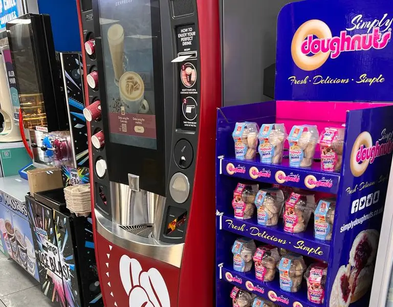 A Simply Doughnuts display stand beside a coffee machine on a filling station forecourt